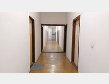 Schmaler Flur in einem einfachen Gebäude mit weiß gestrichenen Wänden und braunen Türrahmen; am Boden ein heller Linoleumbelag. Mehrere Türen führen zu Räumen, und am Ende des Flurs ist ein Schild zu sehen, das nach links zeigt.