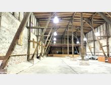 Großzügiger Innenraum einer alten Scheune mit sichtbaren Holzbalken und Fachwerkstruktur. Die Wände bestehen aus unverputztem Backstein, darüber befinden sich mehrere Hängelampen, und der Boden ist aus Beton.