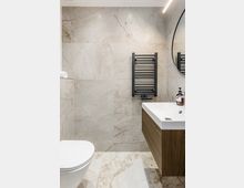 Modernes Badezimmer mit großformatigen, hellgrau marmorierten Wand- und Bodenfliesen. Zu sehen sind ein wandhängendes WC, ein Waschtisch mit Holzunterschrank, ein schwarzer Handtuchheizkörper sowie ein runder Spiegel und eine lineare Deckenleuchte.