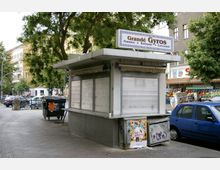Ein geschlossener Imbissstand mit dem Schild „Grandé Gyros“, das Speisen wie Pommes, Bratwurst und Currywurst bewirbt. Der Stand befindet sich an einer Straßenecke mit Bäumen und umliegenden Wohngebäuden; im Hintergrund sind ein Supermarkt und ein Obst- und Gemüsestand zu sehen.