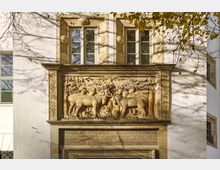 Fassade eines historischen Gebäudes mit detailreichem Sandsteinrelief, das Tiere wie Hirsche und Pferde in einer Waldszene darstellt. Darüber befinden sich zwei Fenster mit steinernen Verzierungen, umrahmt von weißen Wänden und herabhängenden Ästen eines Baumes.