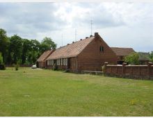 Ein einstöckiges Backsteingebäude mit rotem Ziegeldach befindet sich in einer grünen, offenen Wiesenlandschaft, umgeben von Bäumen und einer niedrigen Backsteinmauer. Im Hintergrund steht ein weißes Auto, und der Himmel ist teils bewölkt.