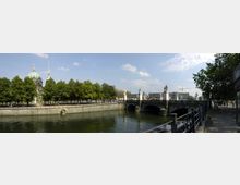 Panoramablick auf den Berliner Dom im Hintergrund, umgeben von Bäumen, mit einem Fluss und der Kronprinzenbrücke im Vordergrund. Die Brücke ist mit Statuen verziert, und der Himmel ist leicht bewölkt.