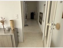 Ein schmaler Flur mit weißen Wänden und hellbeigem Boden, auf der linken Seite ein Metallschrank mit Dekorationen, darunter eine Holzlampe und getrocknete Zweige. Rechts ist eine geöffnete weiße Tür und an der Wand hängt ein kleiner Heizkörper sowie ein Bild. Im Hintergrund steht ein Kleiderständer.