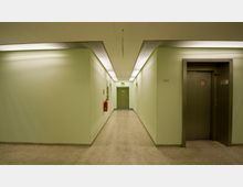 Ein schmaler, hellgrün gestrichener Korridor mit grauem Bodenbelag. Auf der rechten Seite befindet sich ein silberner Aufzug, während auf der linken Wand ein Feuerlöscher hängt; am Ende des Flurs steht eine grüne Tür mit einem Notausgangsschild darüber.