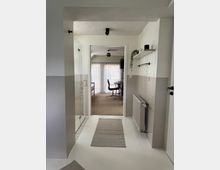 Flur mit weißen Fliesen und hellgrauen Wandfliesen, rechts ein Heizkörper, links eine Glastür zu einer Dusche. Der Flur führt zu einem Wohnzimmer mit Teppichboden, einem Glastisch, schwarzen Stühlen und einem Fenster mit weißen Vorhängen.