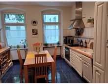 Helle Küche mit blauem Fliesenboden, einem Holzesstisch mit vier Stühlen und großen Fenstern mit weißen Gardinen. Auf der rechten Seite befindet sich eine Einbauküche mit weißen Schränken, einem Herd, einer Abzugshaube und einem Waschbecken.