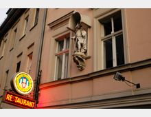 Fassade eines historischen Gebäudes mit einer Fensterfront, einer Statue eines historischen Soldaten unter einem Baldachin und einem leuchtenden Schild mit der Aufschrift „Restaurant“ sowie dem Logo von Budweiser Budvar darunter.