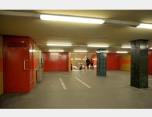 Unterführung mit roten und gelben Wänden, beleuchtet von Deckenlampen. In der Mitte befinden sich blaue Fliesenpfeiler, rechts eine Rolltreppe, und zwei Personen stehen am oberen Ende der Treppe.