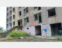 Verlassenes, mehrstöckiges Gebäude mit beschädigten Fenstern und Wänden, die mit Graffiti bedeckt sind. Im Vordergrund wächst wildes Unkraut, und eine Treppe führt zu einem Eingang des Gebäudes.