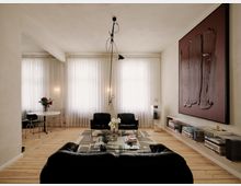 Helles, modernes Wohnzimmer mit breiten Dielenböden, großen Fenstern mit weißen Vorhängen und einer schwarzen Sitzgruppe um einen gläsernen Couchtisch auf einem gemusterten Teppich. Rechts hängt ein großes abstraktes Wandbild über einem niedrigen Regal mit Büchern, links steht ein kleiner runder Esstisch mit Stühlen.