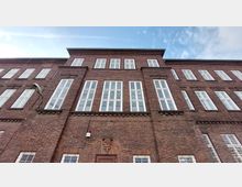 Backsteinfassade eines mehrstöckigen Gebäudes mit großen weißen Sprossenfenstern. Der Gebäudestil erinnert an Industriearchitektur, umgeben von klarem Himmel.