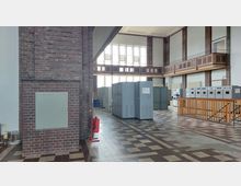 Innenansicht einer industriellen oder technischen Anlage in einem hohen Raum mit roten Klinkerwänden. Der Boden ist mit einem gemusterten Fliesenbelag versehen, und der Raum enthält graue Schaltschränke sowie Geräte an der Wand; eine Holzgalerie läuft entlang der oberen Wand.