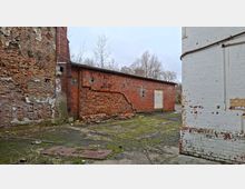Verlassener Industriehof mit verwitterten Backsteinmauern, teils eingestürztem Mauerwerk und Schutt am Boden. Links und hinten sind rote Ziegelgebäude mit beschädigten Wänden zu sehen, rechts eine weiß gestrichene Ziegelwand mit abblätternder Farbe; der Boden ist rissig und von Moos überwachsen.