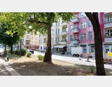 Straßenszene mit mehrstöckigen Wohnhäusern in pastelligen Farben, mit Balkonen und Geschäften im Erdgeschoss. Im Vordergrund befinden sich ein kleiner Park mit Bäumen, Sitzbänken und einem abgestellten E-Roller, während auf der Straße Autos, Fahrräder und eine Baustelle zu sehen sind.