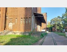 Backstein-Industrielandschaft mit einem alten, mehrstöckigen Gebäude, das kleine Fenster und verblasste Türen aufweist. Im Vordergrund eine Grünfläche, daneben ein asphaltierter Weg mit eingelassenen Schienen, umgeben von Bäumen und weiteren Backsteinbauten.