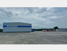 Freifläche mit einer großen Lagerhalle aus Metall mit der Aufschrift „Schwedter Hafen“ und blau-weißen Fassadenelementen. Im Hintergrund sind Betonmauern, rote gestapelte Gegenstände und Bäume unter einem bewölkten Himmel sichtbar.