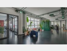 Heller, moderner Bürobereich mit großen Fenstern, Pflanzen in Töpfen und einigen Sitzgelegenheiten, darunter ein blauer Stuhl und ein Sofa mit Kissen. Sichtbar sind grüne Raumteiler mit hängenden Pflanzen und Glaswände, die angrenzende Arbeitsräume zeigen.