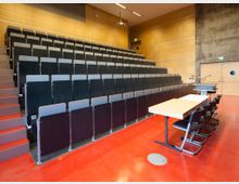 Hörsaal mit ansteigenden Sitzreihen aus Holz und Metall, rotem Boden und einer Dozentenfläche mit Tisch, Stühlen und einem Rednerpult. Die Wände sind teils mit Holzpaneelen verkleidet, eine Tür mit rundem Fenster befindet sich rechts im Hintergrund.