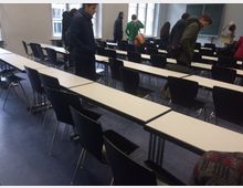 Ein Klassenzimmer mit langen weißen Tischen und schwarzen Stühlen, angeordnet in Reihen. Im Hintergrund befinden sich ein Fenster, Tafeln an der Wand und eine Gruppe von Personen, die das Zimmer verlassen.