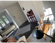 Helles Wohnzimmer mit weiß gestrichenen Wänden, großen Fenstern, einer eingebauten Sitzbank, einem dunklen Fliesenboden und einem Holztisch. Auf einem Teppich steht eine Stehlampe, und ein kleiner Schreibtisch mit Schreibmaschine steht an der Wand.