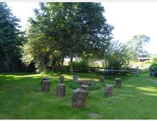 Grüne Gartenlandschaft mit Rasenfläche, auf der mehrere Baumstümpfe um eine Feuerstelle herum arrangiert sind. Im Hintergrund stehen Bäume, Büsche, ein Holzzaun und eine Tischtennisplatte; links und rechts befinden sich weitere Pflanzen und vereinzelte Holzelemente.