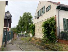 Eine schmale gepflasterte Straße führt an einem Gebäude mit hellem Putz und grünen Fensterläden vorbei, das teilweise mit Natursteinen verkleidet ist. Im Hintergrund ist ein kleiner Kanal mit Ufervegetation und einem Baum zu sehen; links steht eine alte Straßenlaterne neben einem Zaun.
