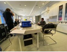 Konferenzraum mit modernen Möbeln, einem großen weißen Tisch in der Mitte, umgeben von mehreren Stühlen. Auf dem Tisch stehen ein Laptop und Dokumente; im Hintergrund sind Personen bei einem Gespräch sowie technische Geräte und weiße Schränke zu sehen.