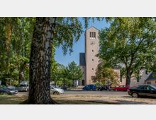 Kirchplatz mit einer modernen Kirche im Hintergrund, die einen rechteckigen Glockenturm mit Uhr und Spitzdach hat. Im Vordergrund steht ein Baum, umgeben von parkenden Autos und Bäumen, die den Bereich säumen.