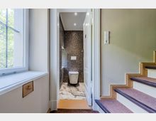 Blick vom Treppenhausflur in ein kleines, modernes Gäste-WC mit dunkler Mosaikfliesenwand, wandhängendem WC und hellem Steinboden. Links befindet sich ein großes Fenster, rechts eine Treppe mit Teppichläufer und Holzstufenkanten.