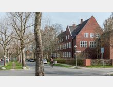 Rote Backsteingebäude mit Satteldach und großen weißen Fenstern, gelegen an einer baumgesäumten Straße mit parkenden Autos; im Vordergrund fährt eine Person mit dem Fahrrad.