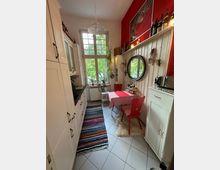 Schmale Küche mit weißer Einbauküche auf der linken Seite und einem Holztisch mit rot-kariertem Tuch und roten Stühlen auf der rechten Seite. Großes Fenster mit Sprossen, bunte Teppiche auf dem Boden, rote Akzentwand mit Dekorationen und Spiegel, sowie Pflanzen und Flaschen als Dekoelemente.