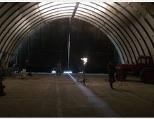 Ein großes, gewölbtes Industriegebäude mit trapezförmigem Dach aus Metallstreben. Im Inneren stehen mehrere Personen und ein roter Traktor, während Scheinwerfer und Kameraausrüstung offenbar für Filmaufnahmen oder eine Produktion eingerichtet sind.