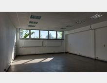 Leerer Raum mit weißen Wänden, einer abgehängten Decke mit Rasterleuchten und grauem Boden. An der Stirnwand befinden sich Fenster in der oberen Hälfte, darunter weiße Heizkörper.