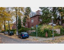Straßenszene in einem Wohnviertel mit herbstlicher Stimmung, flankiert von roten Backsteinhäusern mit Satteldächern und Bäumen, deren gelbes Laub auf dem Boden liegt. Am Straßenrand parken mehrere Autos, und ein Straßenschild zeigt die Kreuzung von Hartmannstraße und Devießenweg an.