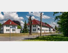 Straßenansicht mit weißen, zweistöckigen Wohnhäusern im Landhausstil, die grüne Fensterläden und rote Ziegeldächer haben. Im Vordergrund ist eine Straße mit Straßenschildern und einem baumlosen Stamm, umgeben von Rasenflächen und Bäumen.