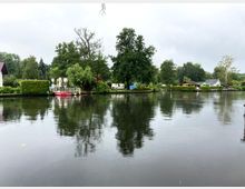 Flussufer mit ruhiger Wasseroberfläche, umgeben von Bäumen, grünen Hecken und kleinen Häusern sowie Bootsanlegern; im Hintergrund ein rotes Boot am Ufer.