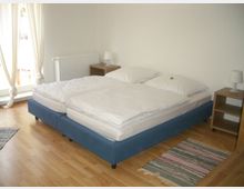 Schlafzimmer mit hellem Holzboden, einem Doppelbett mit blauem Bettgestell und weißer Bettwäsche. Links im Hintergrund befinden sich ein großes Fenster mit langen, hellen Vorhängen und ein Heizkörper, daneben zwei Nachttische mit Lampen. Auf dem Boden liegen kleine, gestreifte Teppiche.