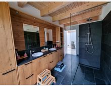Modernes Badezimmer mit Sichtdachstuhl aus Holz. Links eine große Schrank- und Waschtischkombination mit zwei Waschbecken und Spiegeln, rechts eine bodenebene Dusche mit schwarzer Verkleidung und Regenduschkopf.