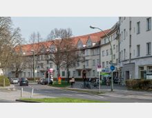 Wohngebiet mit mehrstöckigen Gebäuden im Stil früherer Jahrzehnte, gekennzeichnet durch rote Ziegeldächer und helle Fassaden. Im Vordergrund eine Straße mit Autos, Fahrrädern und Fußgängern sowie ein kleiner Bereich mit Geschäften unter Arkaden.