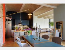 Wohnraum in einem modernen Loft mit offenem Design: Im Vordergrund ein Esstisch mit blauer Oberfläche, darauf Kerzenhalter und eine Vase mit lila Tulpen. Im Hintergrund eine offene Küche mit blauen Wandschränken und Edelstahl-Kühlschrank sowie ein Wohnbereich mit heller Couch und Pflanzen; Decke aus hellen Ziegeln mit Gewölbe-Struktur.