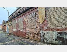 Seitenansicht eines alten Backsteingebäudes mit stark verwitterter Fassade, abgeplatztem Putz und teils ausgebesserten Mauerflächen. Entlang der Wand verläuft ein schmaler Gehweg; am Rand stehen eine Straßenlampe und im Hintergrund weitere Gebäude.