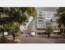 Straßenszene in einer Stadt mit mehrstöckigem Wohn- und Geschäftsgebäude im Hintergrund, das eine Fassade aus Glas und Beton hat. Im Vordergrund gibt es mehrere Autos, einen Fußgänger, einen Radfahrer und Ampeln, umgeben von Bäumen und Gehwegen.