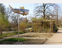 Eine metallische Fischskulptur auf einem hohen Pfosten in einem Park mit gepflastertem Weg, schmiedeeisernem Zaun und Laub abwerfenden Bäumen im Hintergrund.