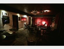 Dimly lit small performance venue with wooden floors, café-style tables and chairs, and a low stage backed by red curtains. A chandelier hangs from the ceiling, and the stage area includes microphones, an acoustic guitar, and an upright piano.