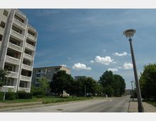 Wohnsiedlung mit mehrstöckigen Plattenbauten, umgeben von Bäumen und Grünflächen. Eine asphaltierte Straße mit Gehweg und Straßenlaternen führt durch die Szenerie, unter einem blauen Himmel mit Wolken.
