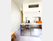 Kleine moderne Küche mit einer Arbeitsplatte aus hellem Material, darunter spiegelnde Schranktüren. Eine Spüle mit metallischem Wasserhahn und eine orangefarbene Brotbox sind sichtbar. Ein lichtdurchfluteter Raum mit schlichtem Design und heller Einrichtung.