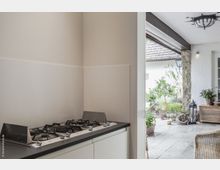 Moderner Küchenbereich mit einem Gaskochfeld und weißem Schrank unter einer hellen Arbeitsfläche, angrenzend an eine überdachte Terrasse mit Steinboden, Pflanzen und rustikalen Möbeln. Im Hintergrund sind eine Hauswand sowie ein Laternen-Lampenschirm sichtbar.