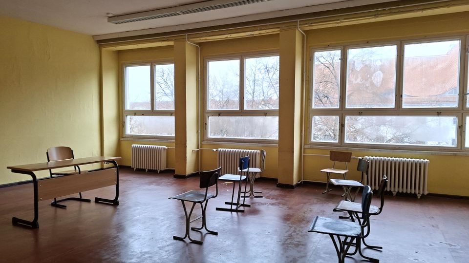 Leerer Klassenraum mit gelb gestrichenen Wänden, großen Fensterfronten und Heizkörpern darunter. Im Raum stehen vereinzelt Schulstühle und Tische auf einem braunen Boden; durch die Fenster sind Bäume und Dächer zu sehen.