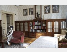 Historischer Salon mit Holzmöbeln, einschließlich einer Bücherwand mit verglasten Türen, einem roten Polstersofa, und einem goldfarbenen Sessel. Eine Schaufensterpuppe in einem Kleid sitzt auf dem Sofa, und an der Wand hängen gerahmte Fotografien und ein Spiegel.